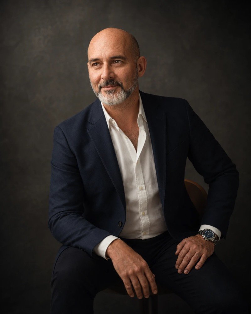 Portrait of Jorge Alexandre Pereira, seated, wearing a dark blue jacket and white shirt, against a dark background.