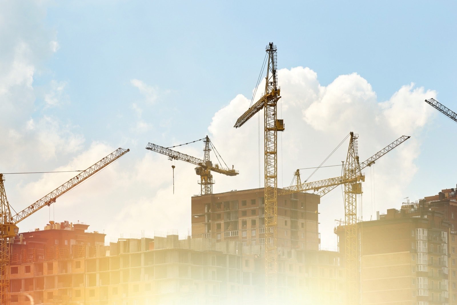 Várias gruas de torre sobre edifícios em construção, contra o céu com sol encoberto.