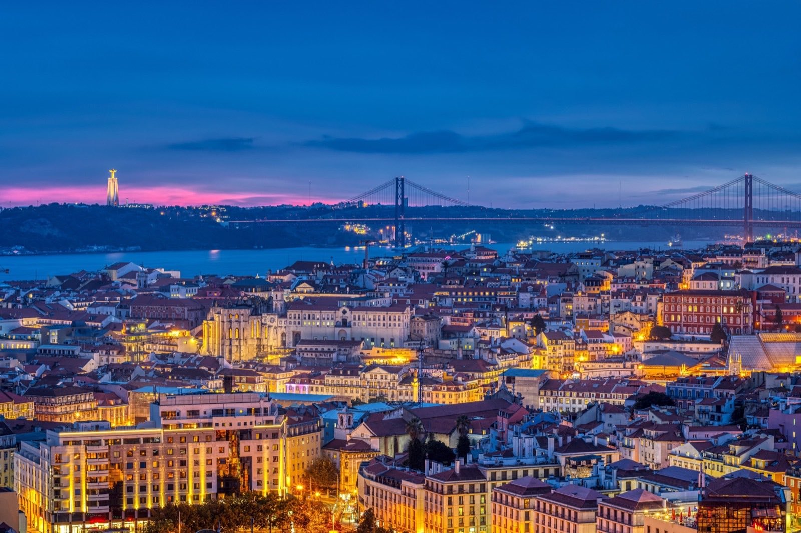 Lisboa ao anoitecer, com as luzes acesas; ao fundo, a Ponte 25 de Abril e o Cristo Rei.