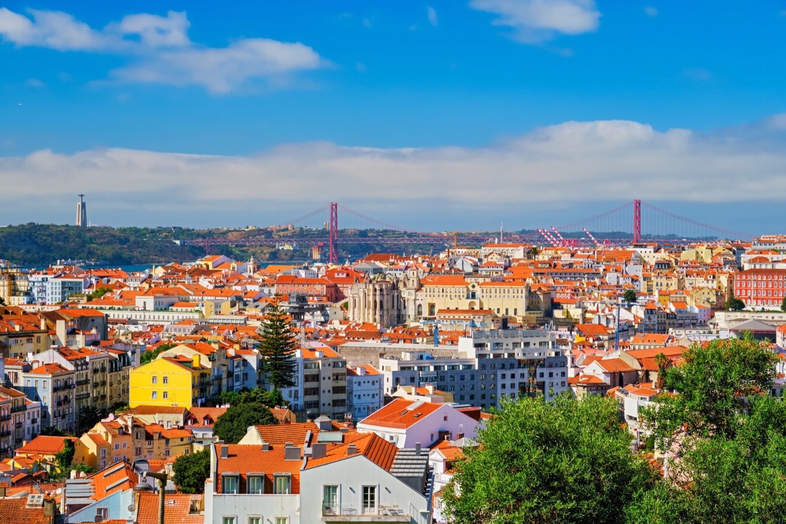 Telhados e prédios de Lisboa densamente agrupados; ao fundo, o Tejo, a Ponte 25 de Abril e o Cristo Rei.